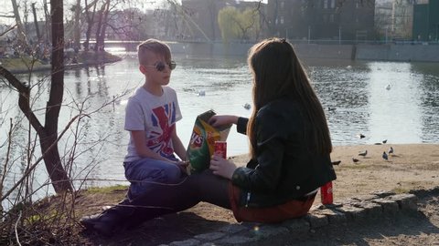 WROCLAW, POLAND - MAR 21, 2019: Teenager young kids friends boy and girl drinking cola can the carbonated beverage and eat crispy potato chips during walking in city park