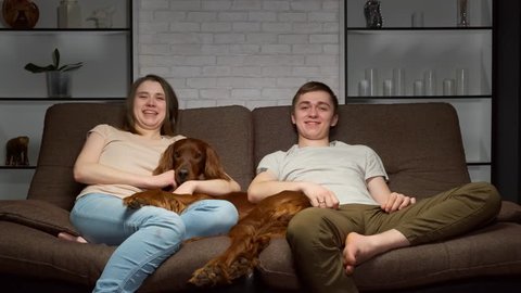 Happy young couple watching tv together with pet sitting on sofa at home.
