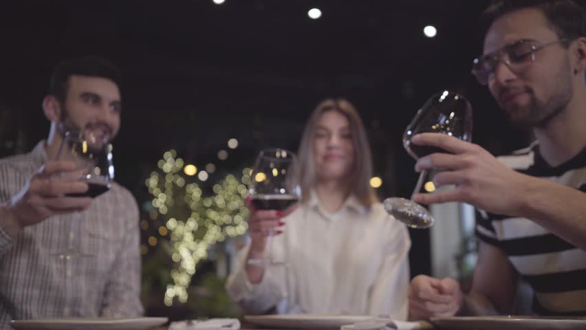 Two men and woman clinking their glases with red wine sitting at the table in modern turkish restaurant. Friends have meeting, relaxing together.