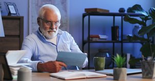 Senior Caucasian man in glasses working late at night on the tablet device in his cabinet at home while working and drinking tea or coffee.