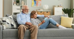 Caucasian small teen girl with long fair hair resting on the sofa with her grandpa in glasses and demonstrating him something on the tablet screen in the living room.