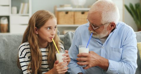 Portrait of the cheerful granfather in glasses and cute granddaughter drinking milk or milkshakes with straws on the couch in the living room.