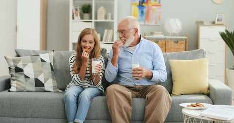 Pretty blonde teen girl sitting together with her grandfather with grey haired on the sofa at home and drinking milk with cookies.