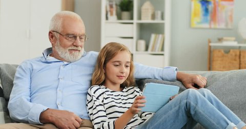 Portrait shot of the excited caucasian grandfather and granddaughter watching video on the tablet device while sitting together on the sofa in the cozy living room.