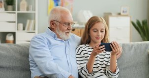 Portrait shot of the Caucasian grandfather and granddaughter resting on the couch in the living room and girl showing something to the grandpa on the phone screen.