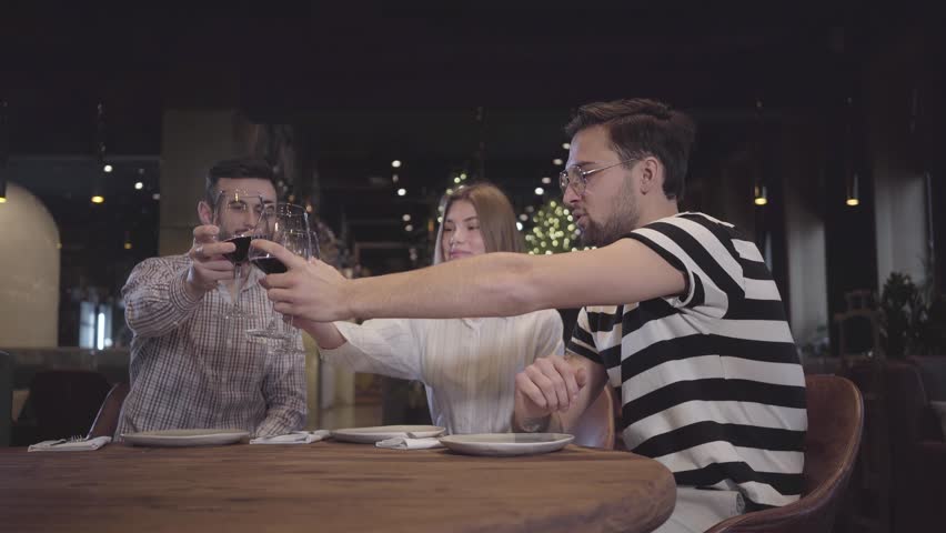 Two men and woman clinking their glases with red wine sitting at the table in modern turkish restaurant. Friends have meeting, relaxing together.