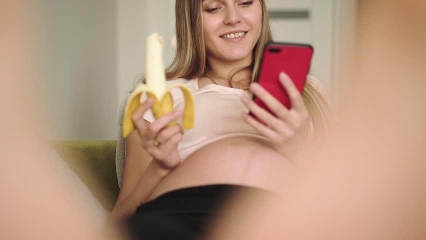 Happy pregnant rests on sofa, eats a banana and uses phone. 4K