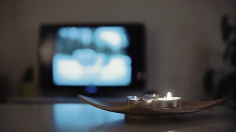 4K stationary shot of tealight candle being blown out on a wooden board. Blue smoke. Winter mood with a television in background.