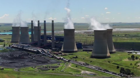 Ominous shadow of dark cloud falls over coal power station in South Africa. Orbit aerial view of power plant, coal stockpile, green summer landscape, smoke rising from six cooling tower chimneys