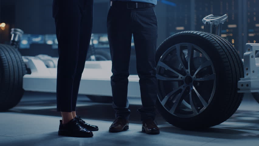 Male and Female Automobile Design Electric Car Engineers Having Conversation, Use Clipboard to Check Maintenance Issues. Innovative Hybrid Vehicle Platform Chassis Concept with Wheels, Engine, Battery