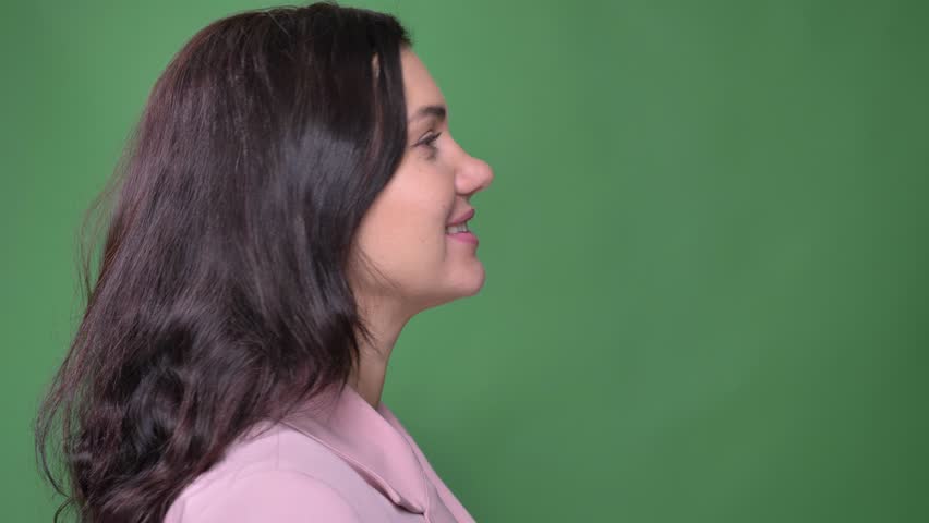 Young brunette businesswoman in pink jacket turns to camera and watches delightedly into camera on green background.