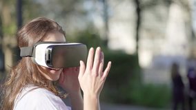 Pretty serious Caucasian woman with long curly hair in blue shirt wearing virtual reality glasses in park, managing new reality with hands, smiling. High technology, VR concept