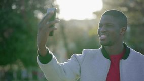Young Afro-American short-cut muscular man in rose T-shirt and grey jacket having video chat on phone in park, showing surroundings. Communication, lifestyle concept