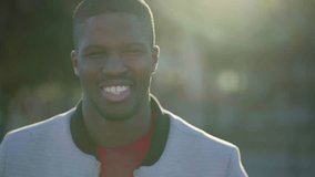 Medium shot of young Afro-American short-cut muscular man in rose T-shirt and grey jacket standing in park, looking at camera, smiling. Lifestyle concept
