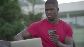 Medium shot of young Afro-American short-cut muscular man in rose T-shirt sitting on bench in park typing on laptop, paying online with credit card. Work, online shopping concept
