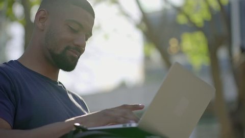Bottom view of young Afro-American short-cut muscular man with stylish beard in blue T-shirt sitting on bench in park, working on laptop. Sun shining. Lifestyle, work concept 