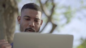Bottom view of young Afro-American short-cut muscular man with stylish beard in blue T-shirt working on laptop, receiving good results, exulting. Close-up short. Success, work concept 
