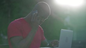 Medium shot of young Afro-American short-cut muscular man in roseT-shirt sitting on bench in park, typing on laptop, talking on phone. Side view. Sun shining. Lifestyle, work concept 