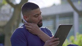 Side view of young Afro-American short-cut muscular man with stylish beard in blue T-shirt sitting on bench in park, talking on phone, searching information on tablet. Communication, lifestyle concept