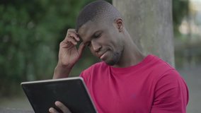 Medium shot of young Afro-American short-cut muscular man in rose T-shirt sitting on bench in park, watching football match on tablet, feeling disappointed, massaging head. Lifestyle, sport concept 