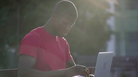 Medium shot of young Afro-American short-cut muscular man in roseT-shirt sitting on bench in park, working on laptop, typing. Side view. Sun shining. Lifestyle, work concept 