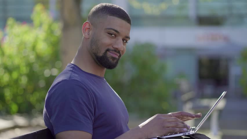 Side view of young Afro-American short-cut muscular man with stylish beard in blue T-shirt sitting on bench in park, working on laptop, turning head to camera. Sun shining. Lifestyle, work concept 