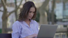 Pretty Caucasian woman with long curly hair in blue shirt sitting in park, working on laptop. Side view. Work, lifestyle concept