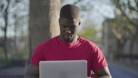 Medium shot of young Afro-American short-cut muscular man in roseT-shirt sitting on bench in park, working on laptop, smiling. Front view. Sun shining. Lifestyle, work concept 