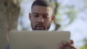 Bottom view of young Afro-American muscular man with stylish beard in blue T-shirt sitting on bench in park, working on laptop, thinking, having headache, massaging temples. Lifestyle, work concept 