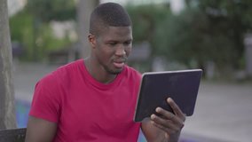 Medium shot of young Afro-American short-cut muscular man in roseT-shirt sitting on bench in park, watching football match on tablet, celebrating goal of his team. Lifestyle, sport concept 