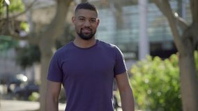 medium shot of young Afro-American short-cut muscular man with stylish beard in blue T-shirt standing in park, looking at camera, smiling. Front view. Lifestyle concept concept 