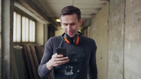 Medium shot of carpenter with protective earmuffs around his neck walking along corridor in industrial building and text messaging on cell phone in slow motion