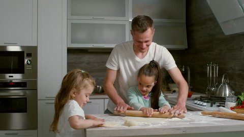 Medium shot of father and two daughters making dough together in kitchen, man teaching one of girls to use rolling pin