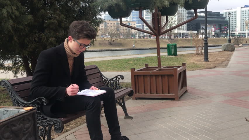 A young man sits on a bench and makes sketches in a notebook. Turns pages. Spring, cool weather, the guy is wearing a black coat. Frame rotation 360 degrees.