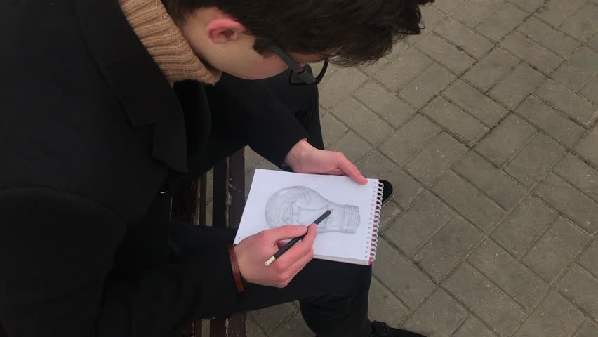A young man sits on a bench and makes sketches in a notebook. Spring, cool weather, the guy is wearing a black coat. Closeup illustration of his work, top view