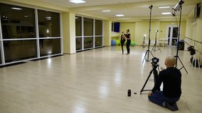Video cameraman filming as two slim cute girls sisters artistic gymnasts in black sportswear make warm-up in gym and perform muscle stretching exercise. Rhythmic gymnastics for child and teenager.