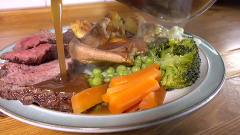 Close POV circular dolly shot of steaming thick gravy being poured all over a Sunday dinner plate on a pine kitchen worktop, of traditional roast beef, vegetables and Yorkshire pudding.