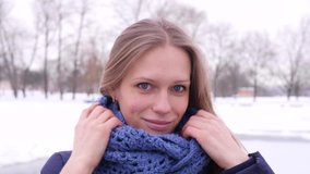 Portrait of a pretty blue-eyed woman in the winter city park who looking at the camera and corrects her long hair and blue knitted scarf in 4K slow motion close up video.