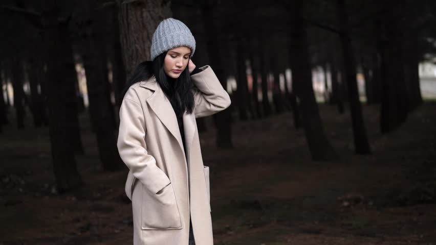 attractive girl in knitted hat and long spring coat walks through old pine wood in morning slow motion