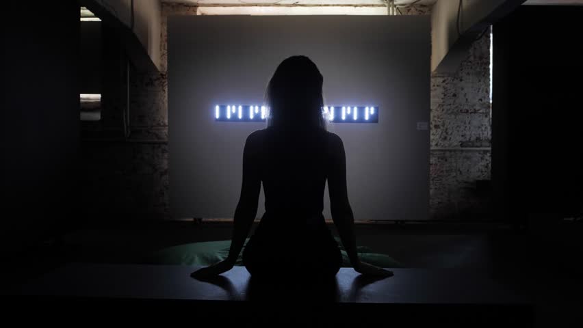 girl with long loose flowing hair silhouette sits in front of display with white numbers in dark room backside view