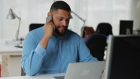 Smiling bearded young man talking on smartphone. Cheerful young brunet businessman talking on phone while sitting at table with laptop. Technology and communication concept