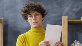 Medium shot of young curly haired woman in glasses and yellow turtleneck showing greeting card template and art supplies used to decorate it while recording video blog