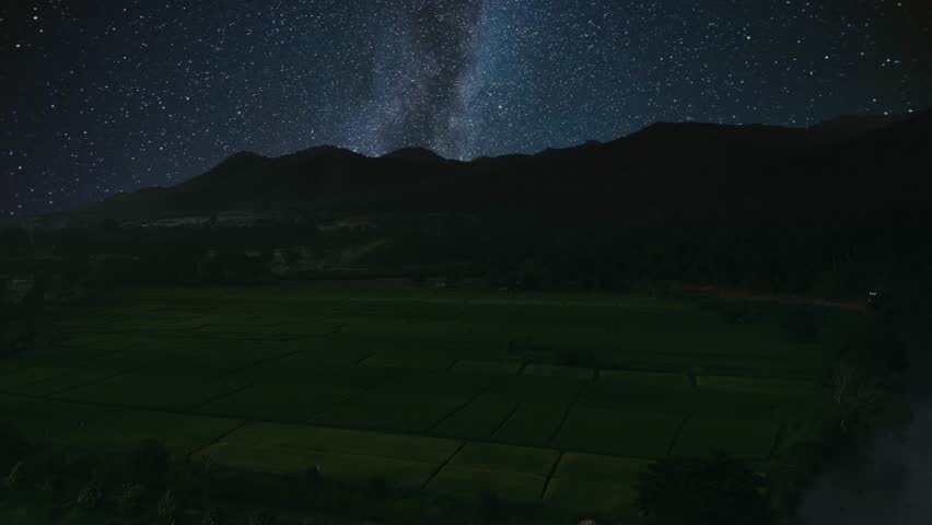Aerial flight above field against mountains with milky way and stars on night sky