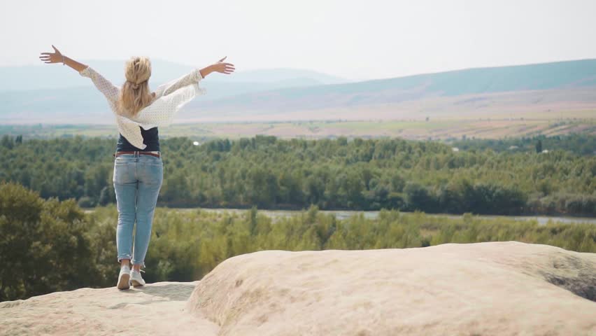 feels flying in wind and flying hair and light summer clothes. lady in jeans and waving white shirt in front of magnificent view of nature from mountain, raising arms upwards, like wings of bird