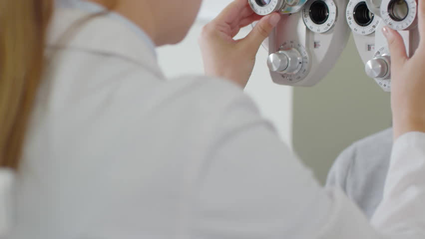 Tilt up shot of female optometrist in white coat changing lenses on phoropter instrument and talking to male patient having eye exam Royalty-Free Stock Footage #1026695471