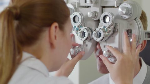 Tilt up shot of female optometrist in white coat changing lenses on phoropter instrument and talking to male patient having eye exam