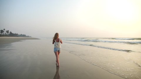 Young attractive girl cute blonde with loose hair goes in sunset rays and taking a photo selfie on a tropical sandy beach on sea shore. Silhouette of slim woman with smartphone in ocean waves.