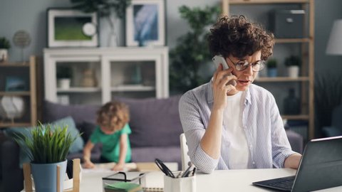 Angry female freelancer is talking on mobile phone and using laptop while her small son is playing in room. Modern business, communication and family concept.