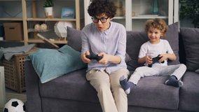 Emotional girl mother is playing video games with little son holding joystick at home on sofa enjoying activity. Indoor fun and modern gadgets concept.