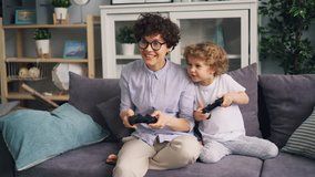 Mother and young child are playing video game on couch at home, pressing buttons on joystick, laughing and having fun. Happy family and modern technology concept.